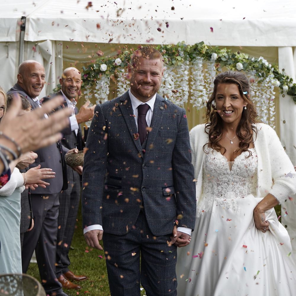 Guests throw confetti over a smiling bride and groom leaving a marquee ceremony