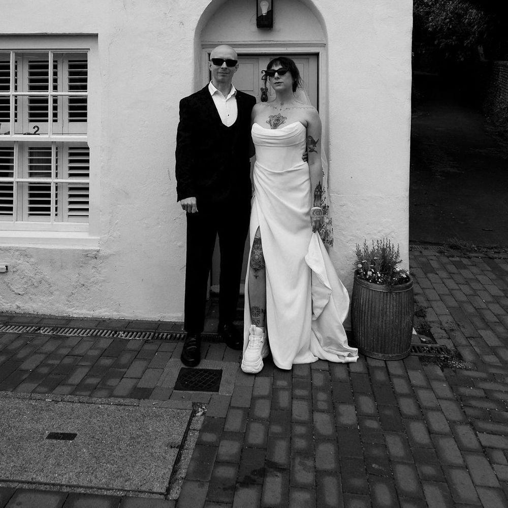 Bride and groom in sunglasses standing by an arched doorway of a white cottage.