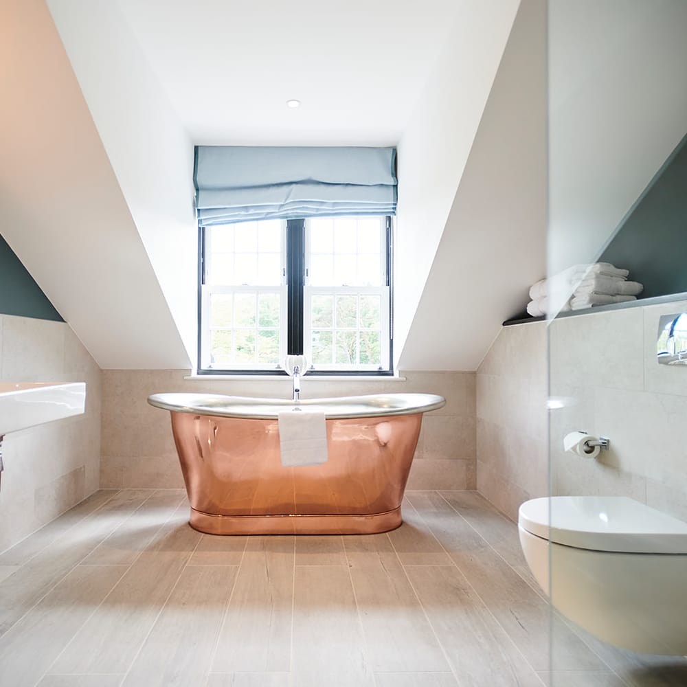 Copper roll-top bath beneath a window in a bathroom with teal walls and neutral tiling