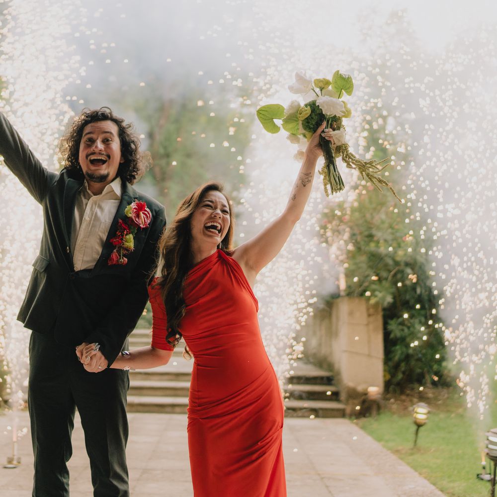 Jubilant couple raise their arms amid sparkler fountains, bride in red dress holding a bouquet