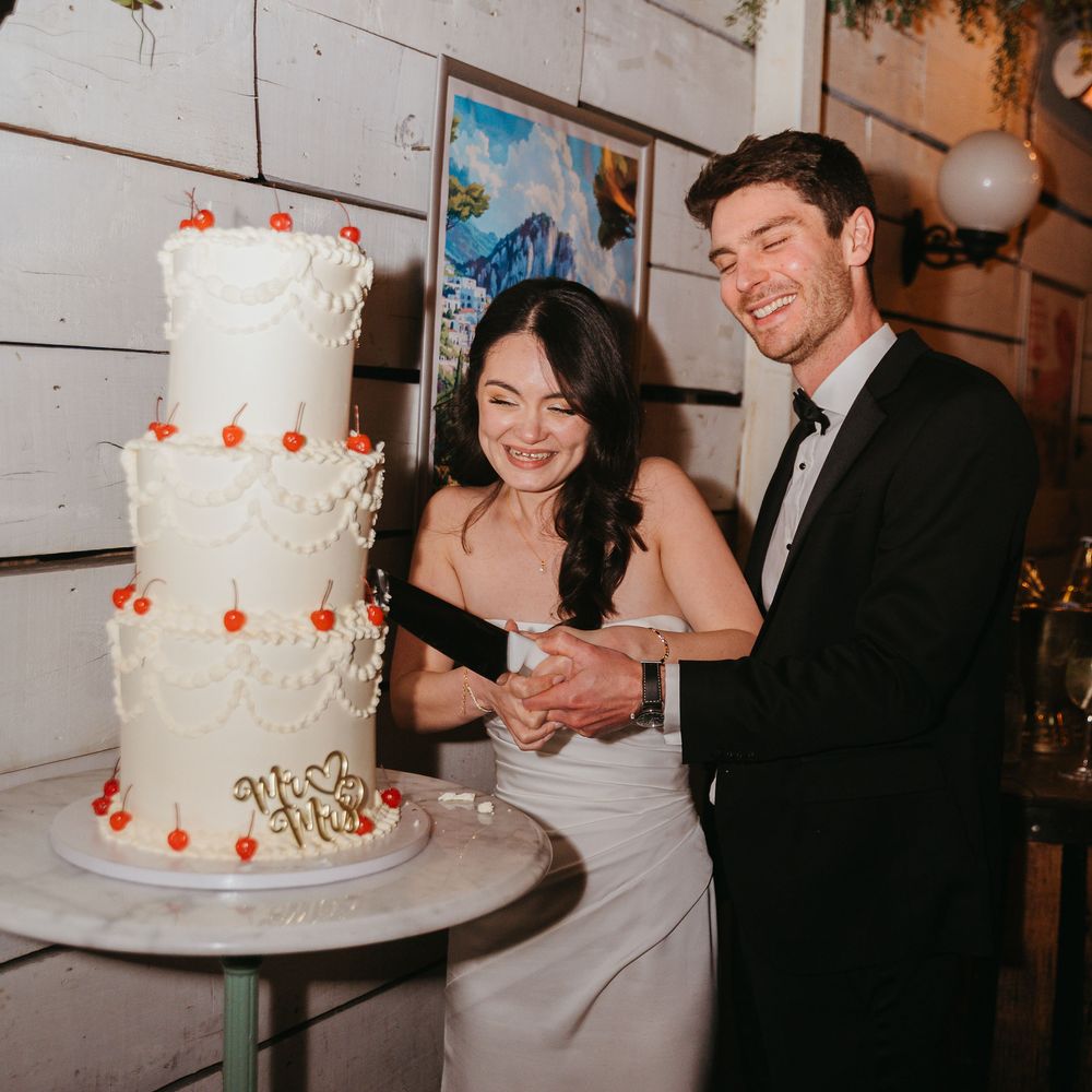 Laughing bride and groom cut a three-tier white cake topped with cherries at their reception