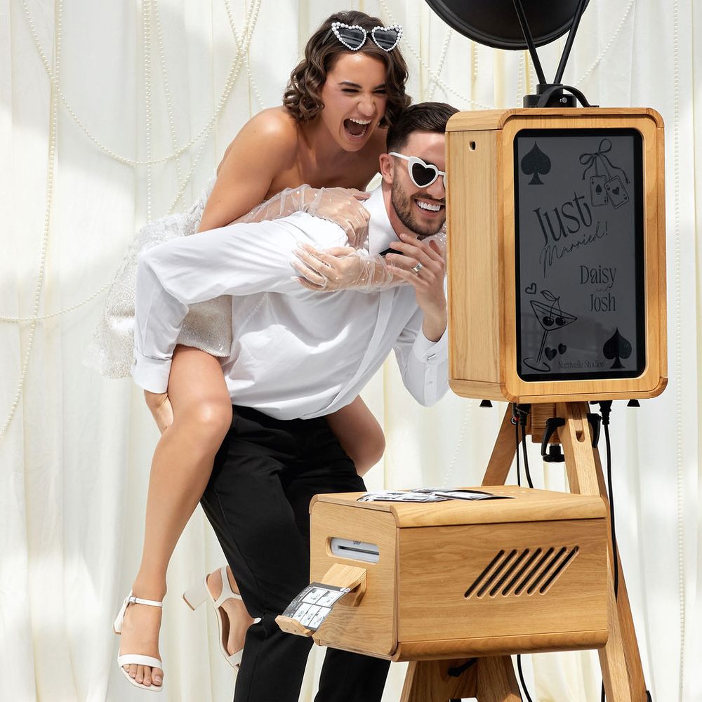Laughing couple in heart-shaped sunglasses pose beside a wooden photo booth against ivory drapes