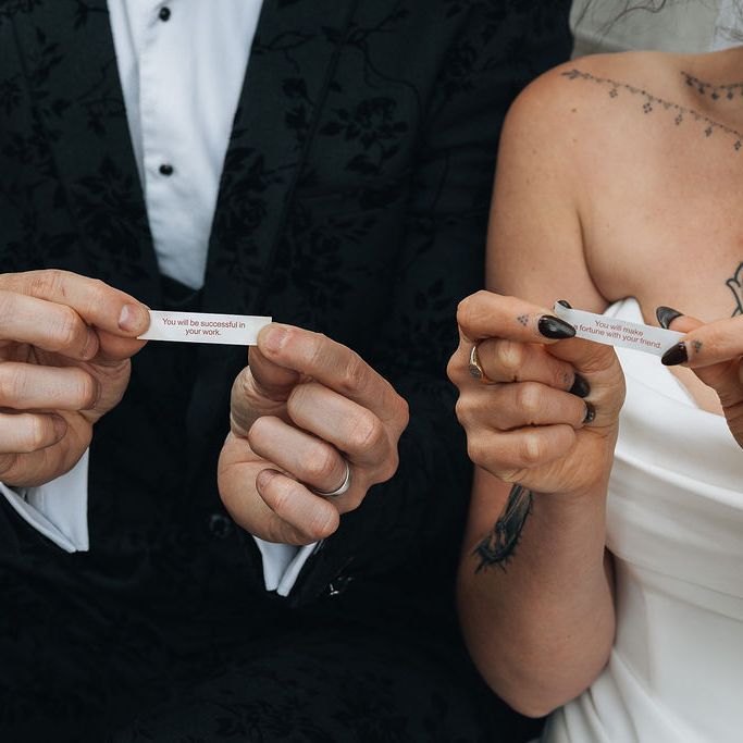 Bride and groom holding fortune cookie messages, with visible tattoos and dark manicure.