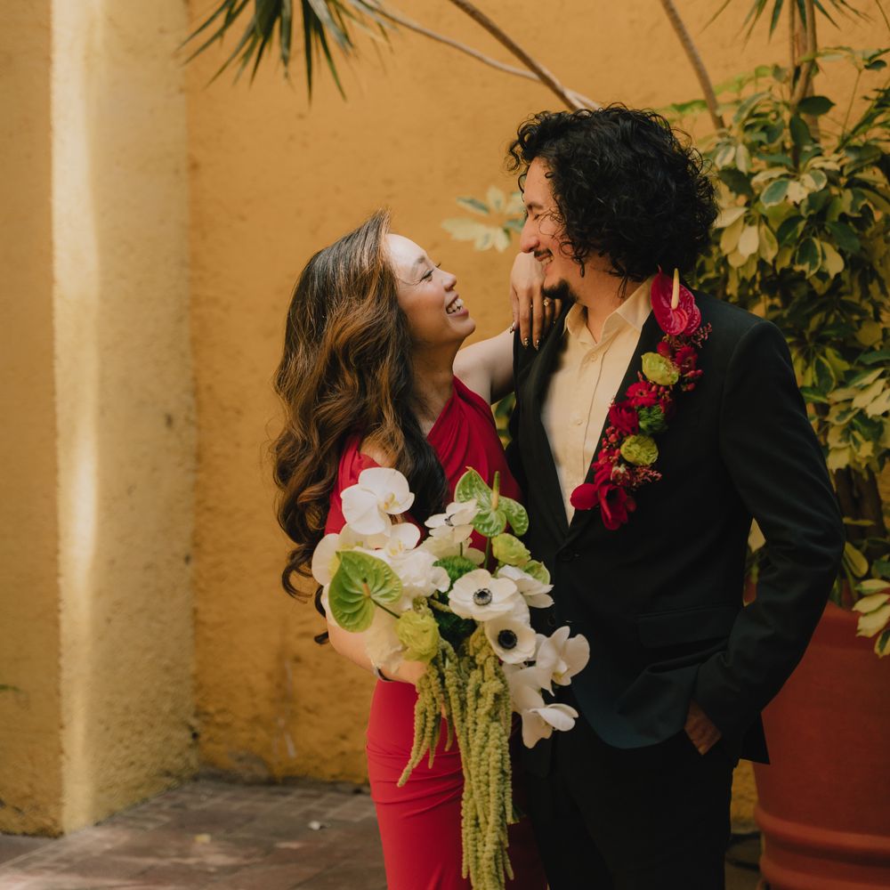 Bride in red dress and groom in black suit share a look beside a sunny yellow wall