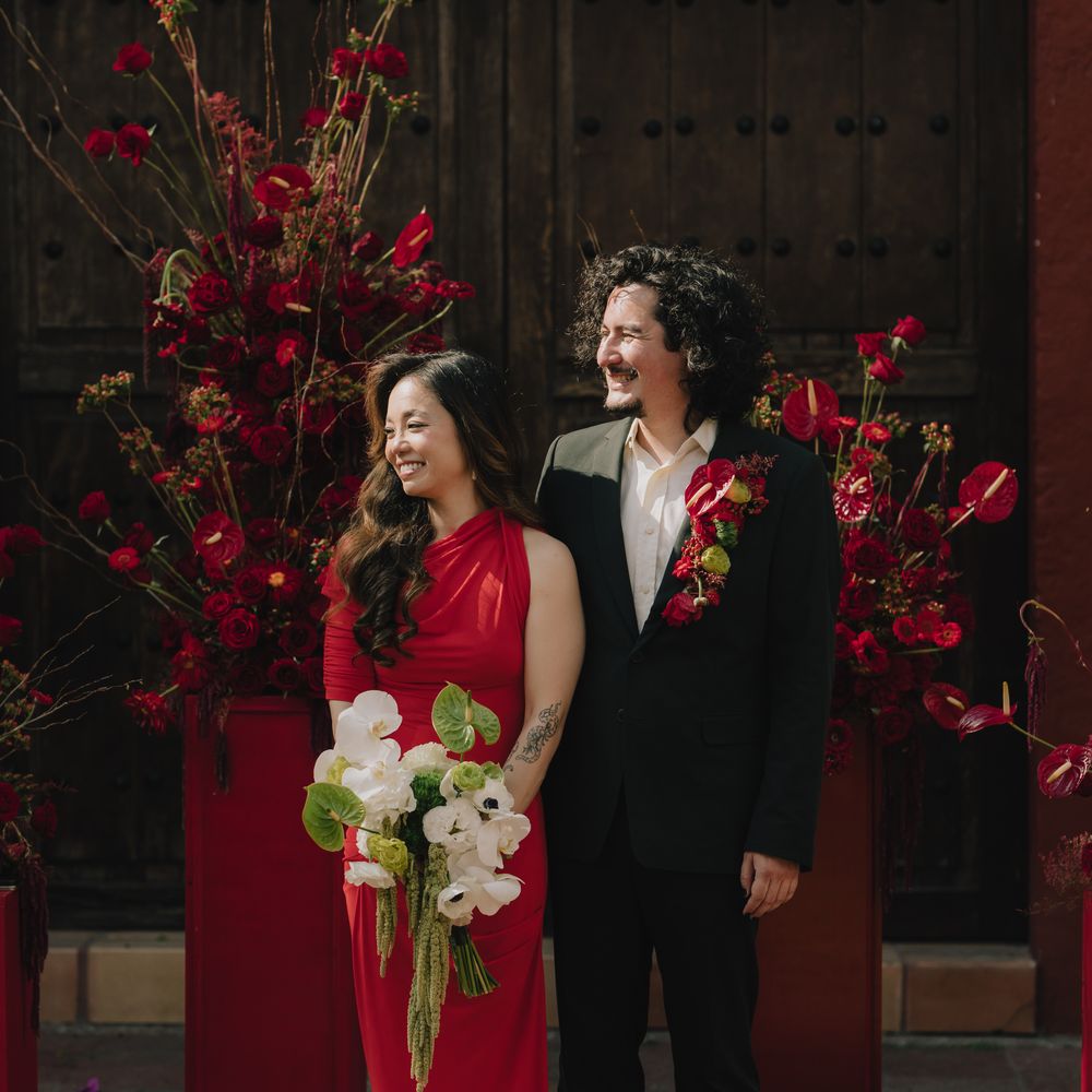 Smiling couple in red gown and black suit stand before dramatic crimson floral displays