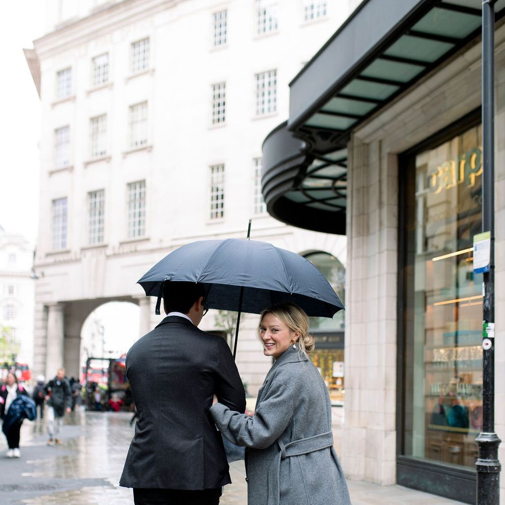 Couple sharing an umbrella on a rainy city street, bride in a grey wool coat glancing back with a smile