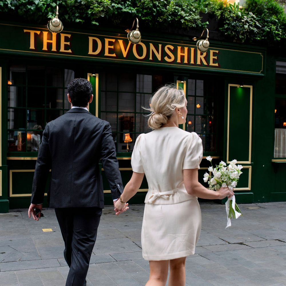 Newlywed couple hold hands walking towards a green London pub with floral facade