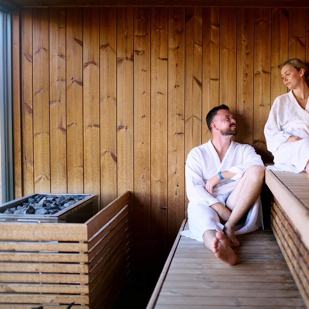 Couple in white robes relaxing together in a warm wooden sauna with heated stones nearby