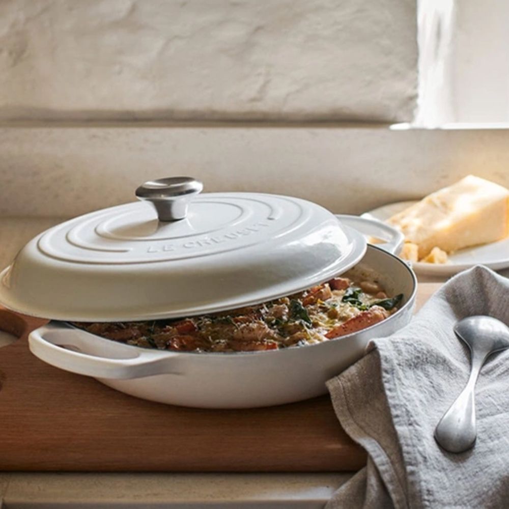 Cream cast iron casserole dish of vegetable bake on a wooden board with bread and cheese