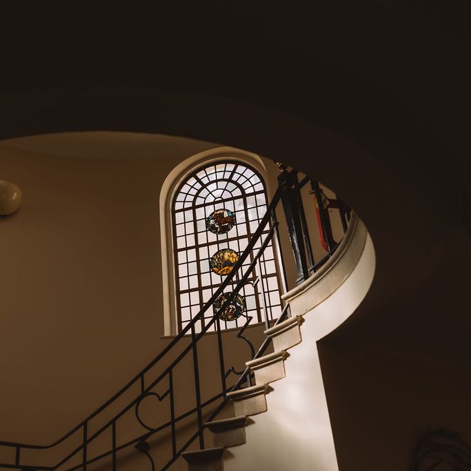 Sweeping curved staircase beneath an arched stained glass window in a dimly lit interior
