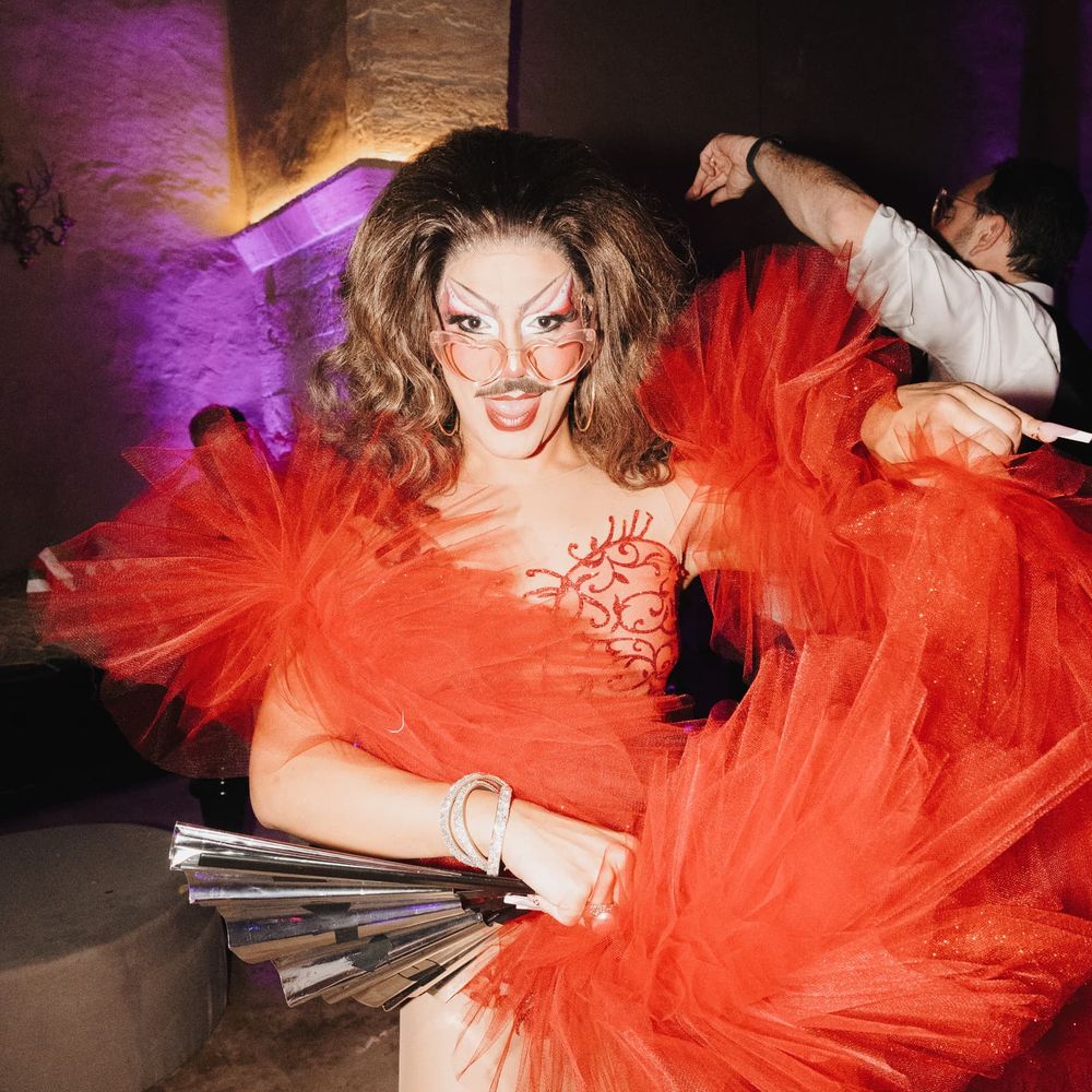 Drag performer in a dramatic red tulle outfit poses under purple lighting at an evening reception