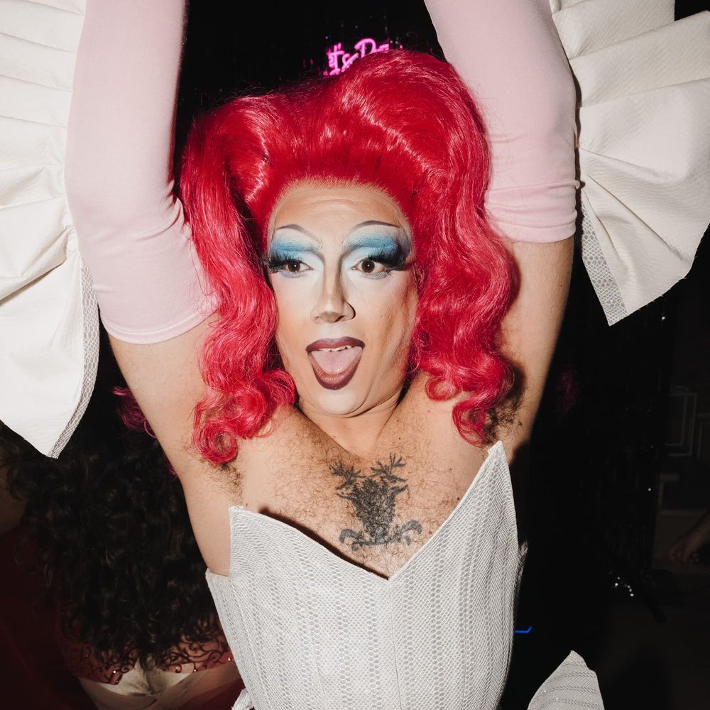 Drag performer in a red wig and structured white corset poses with arms raised on the dancefloor