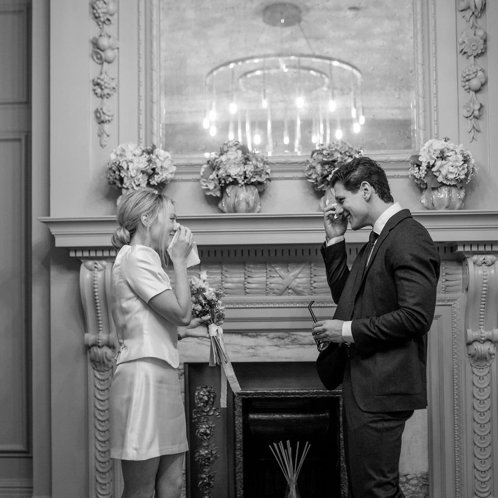 Emotional bride and groom wiping away tears beside an ornate marble fireplace during their ceremony