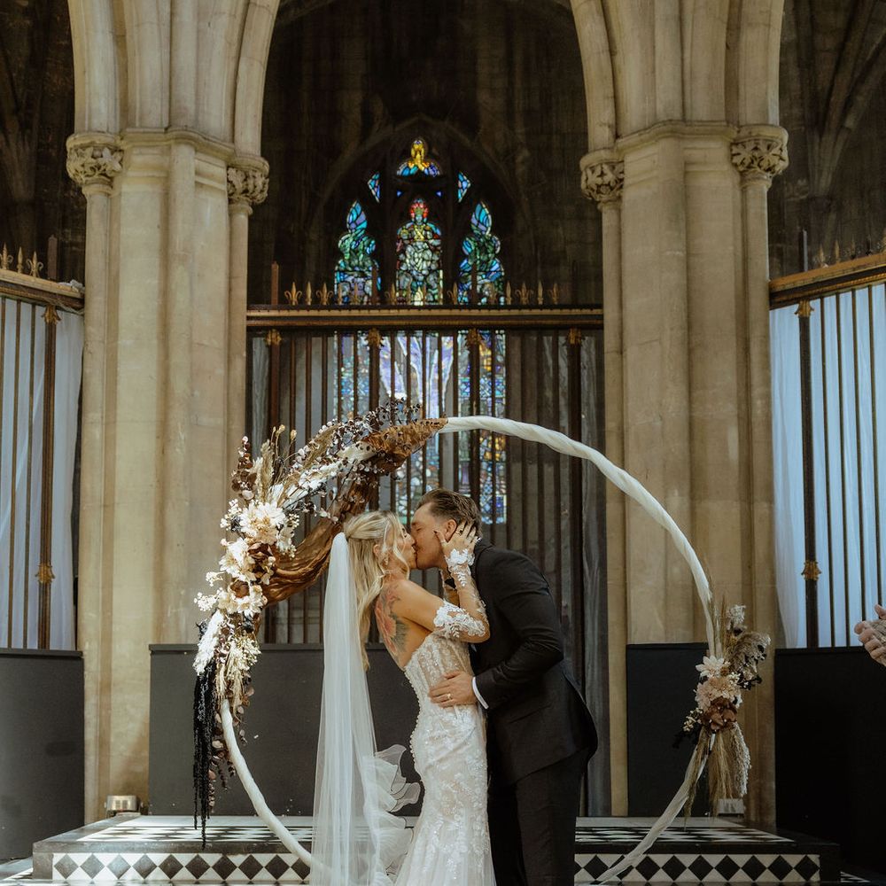 Bride and groom share their first kiss beneath a circular dried floral arch in a Gothic chapel