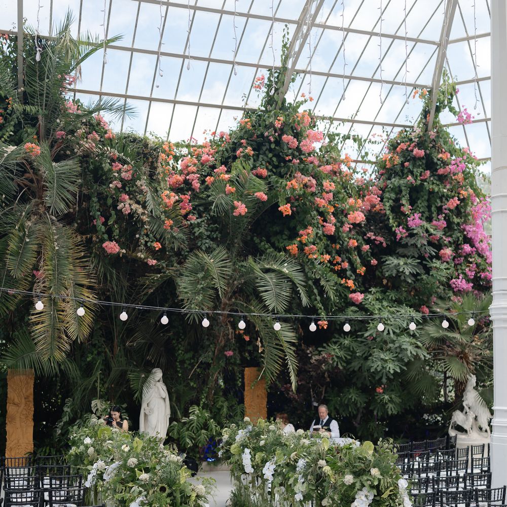 Glasshouse ceremony aisle lined with white blooms and greenery beneath tropical planting and festoon lights