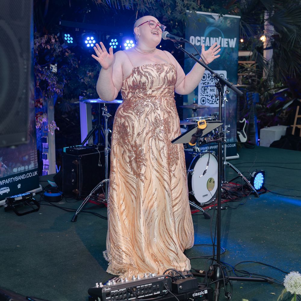 Singer in a gold sequinned gown performs at a microphone on a tropical-styled stage.