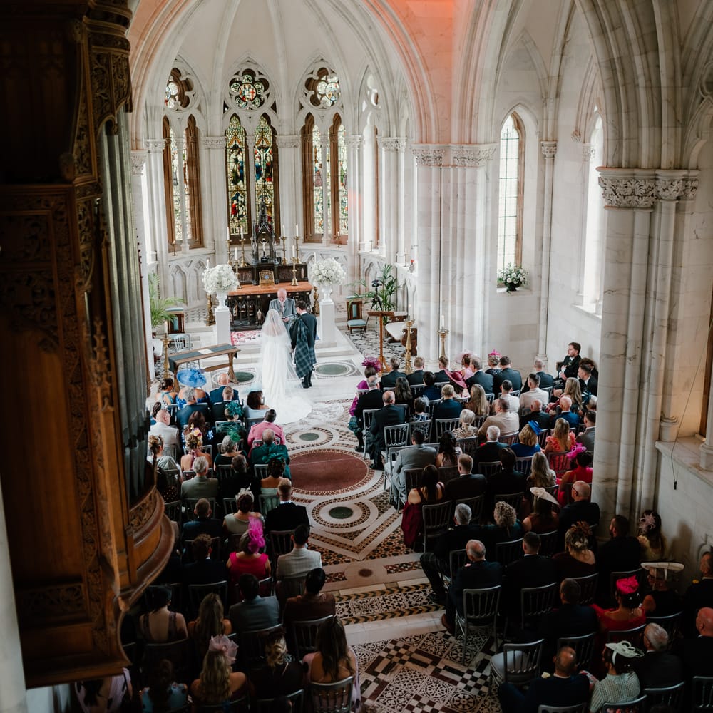 Couple exchange vows at the altar of a grand chapel with stained glass windows and guests seated