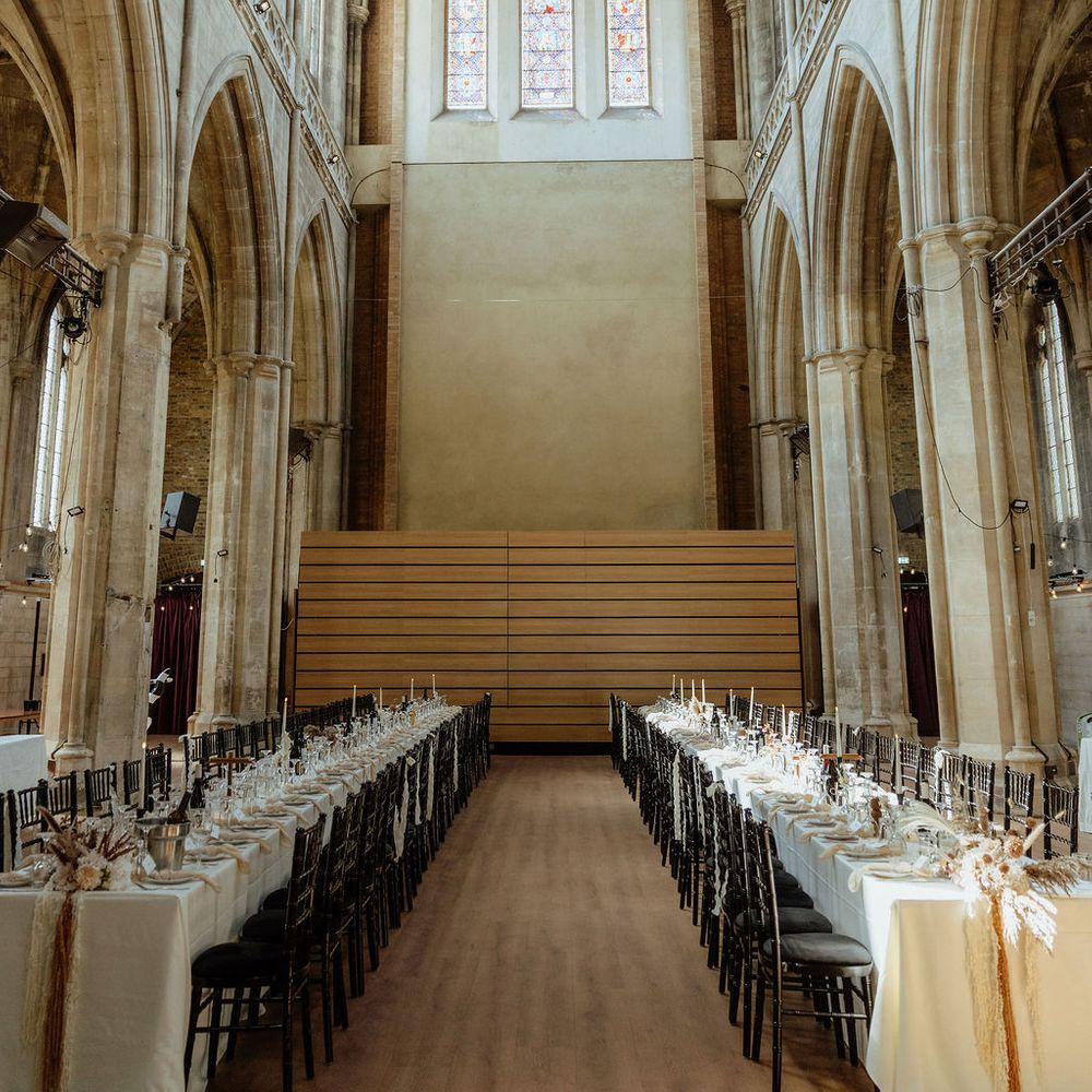 Long banquet tables with pampas grass runners set beneath a stained glass window in a gothic hall