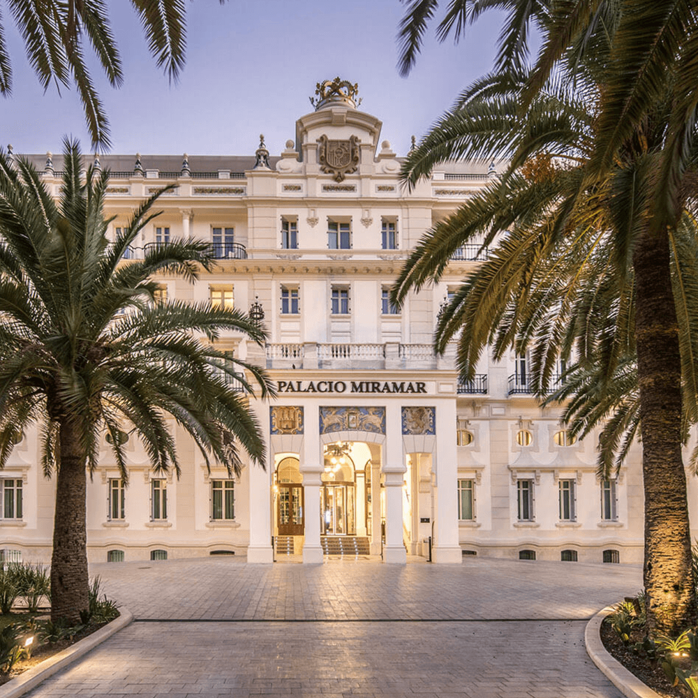 Gran Hotel Miramar Malaga grand white palace hotel facade framed by palm trees at dusk with a lit entrance archway