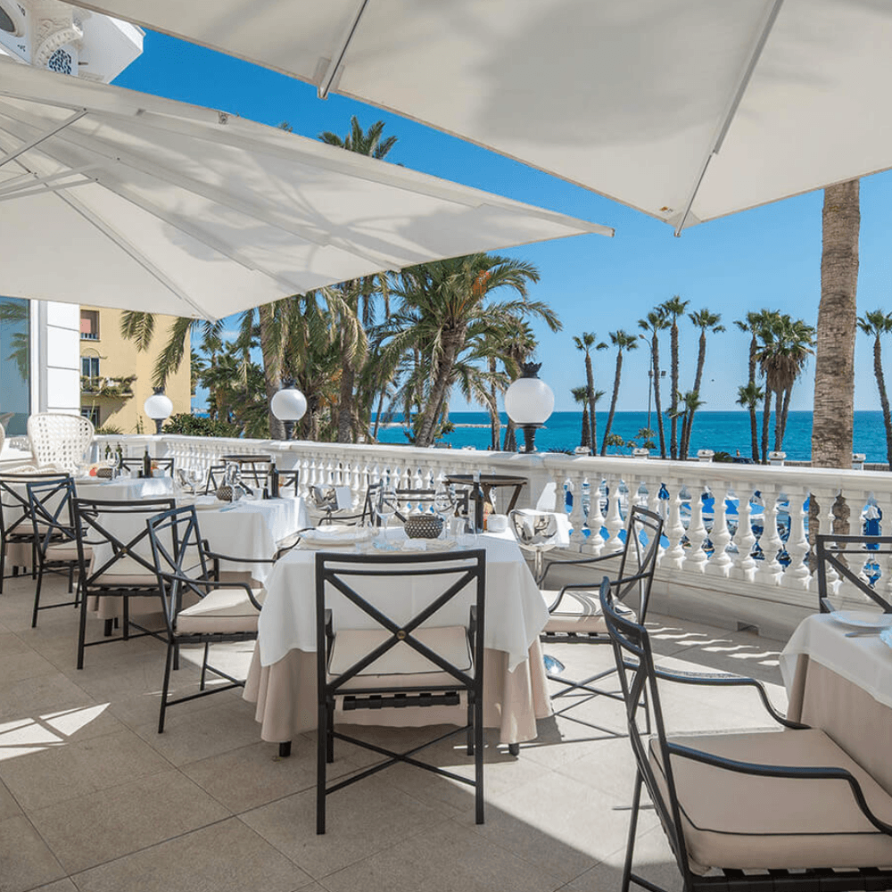 Seaside terrace set with white-linen tables, iron chairs and parasols overlooking palms and the sea