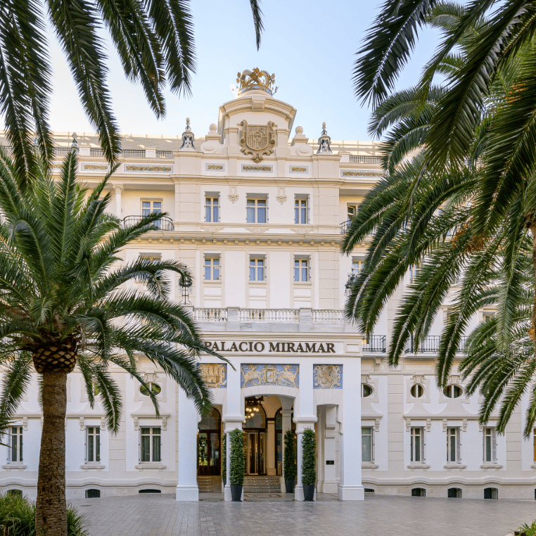 Grand white palace facade framed by palm trees, with ornate crest and tiled entrance archway