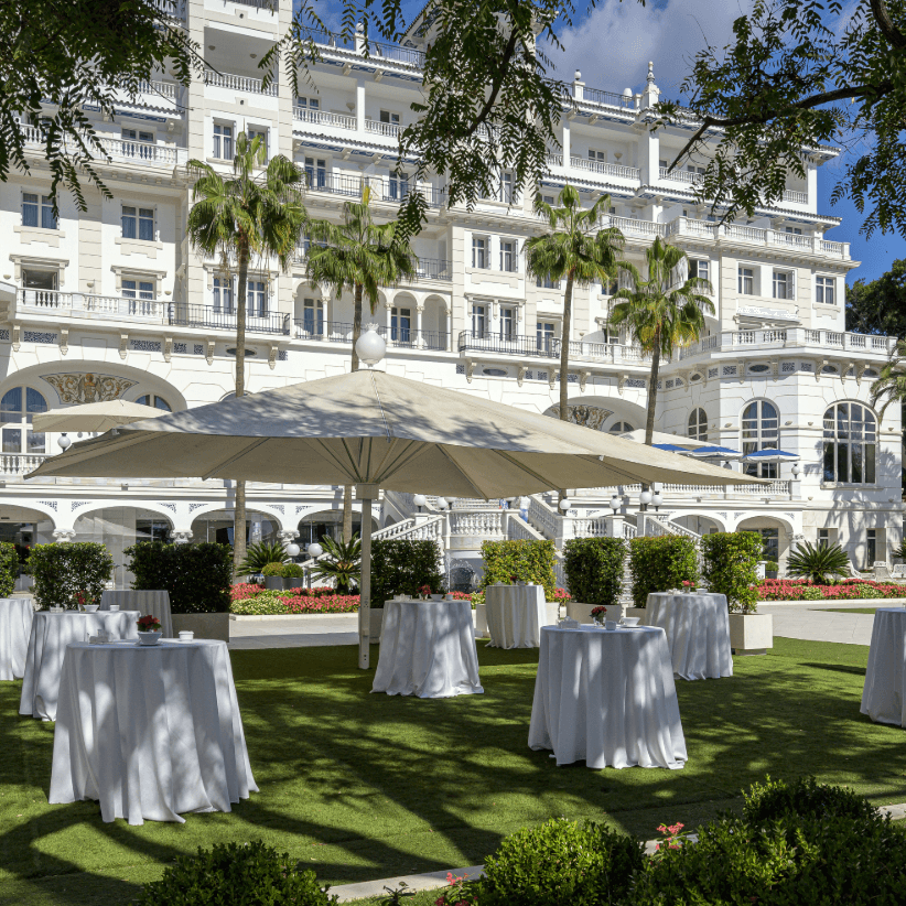 White-clothed cocktail tables on a manicured lawn beside a grand white palace hotel with palms.