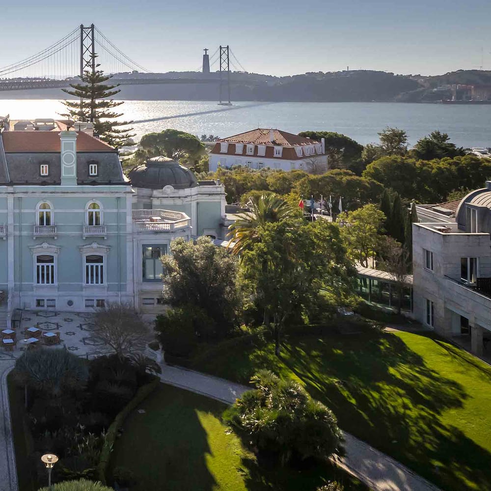 Aerial view of a grand pastel palace with manicured gardens overlooking a river and suspension bridge