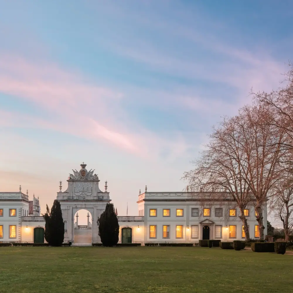Grand white palatial venue with central archway and lit windows at dusk beneath a pink sky