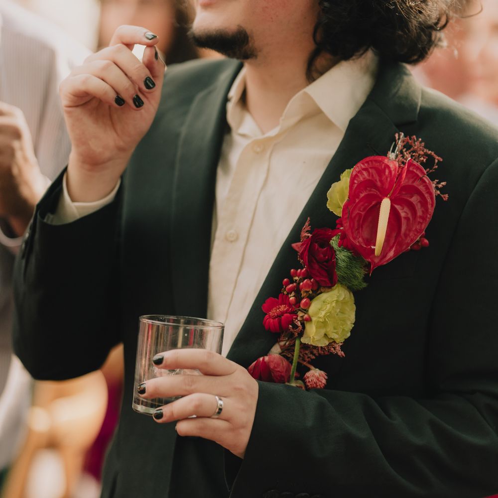 Groom in dark green suit with bold red anthurium buttonhole holding a drink