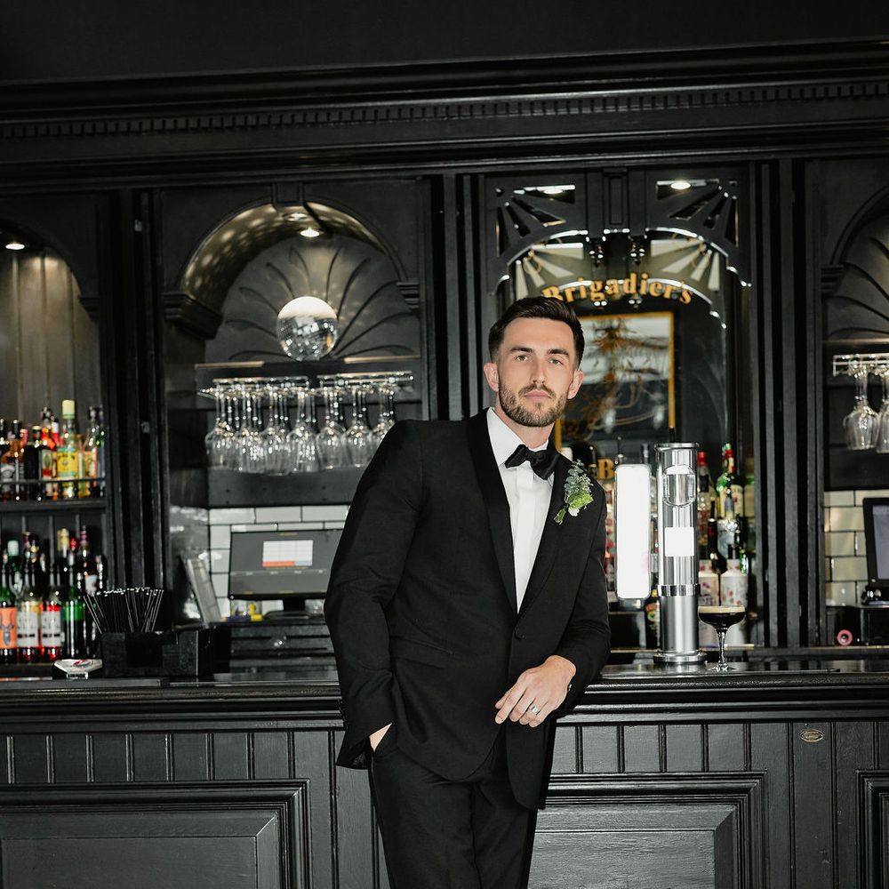 Groom in a black tuxedo and bow tie leaning against a dark bar with a chequered floor