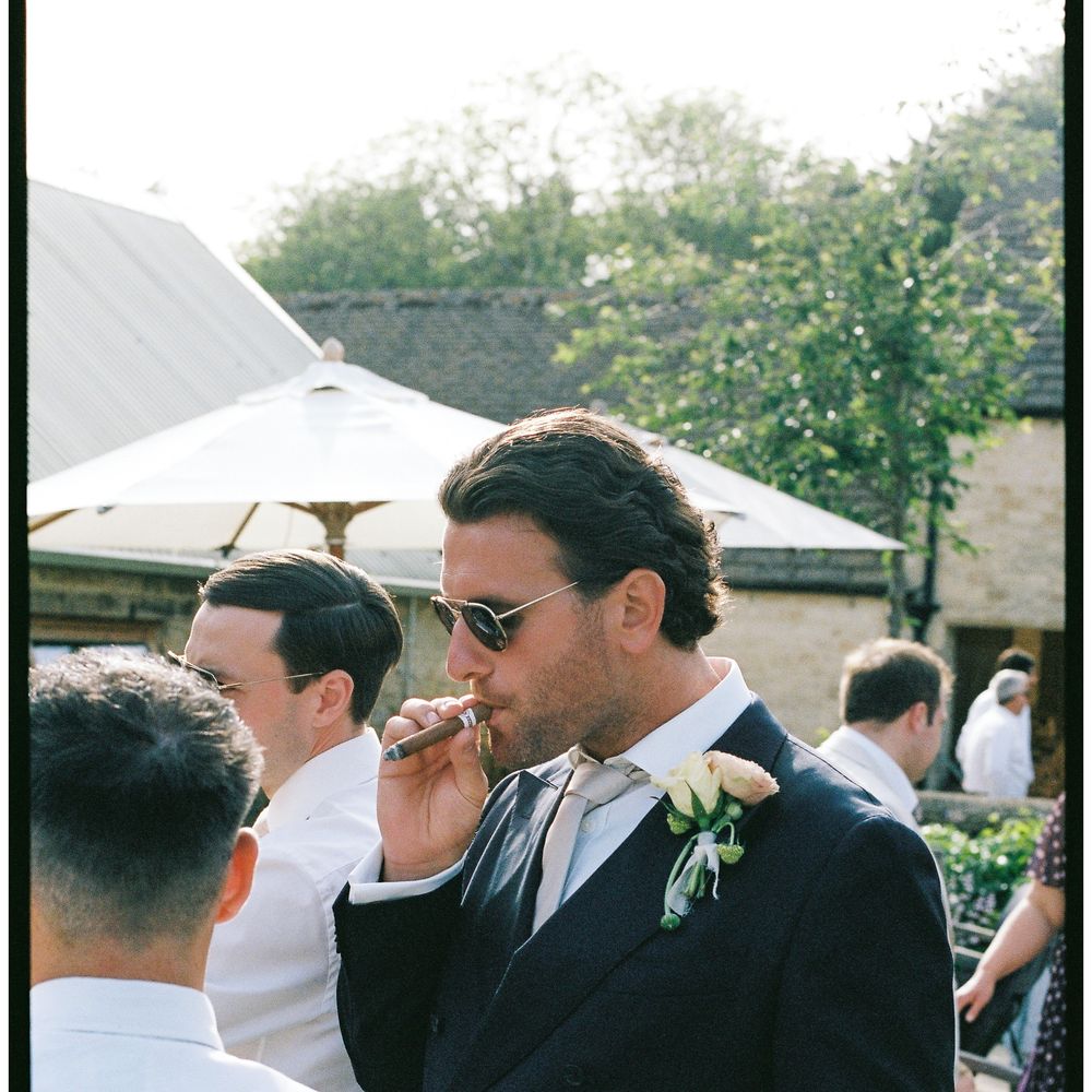 Groom in navy suit and sunglasses smoking a cigar outdoors among guests in sunshine