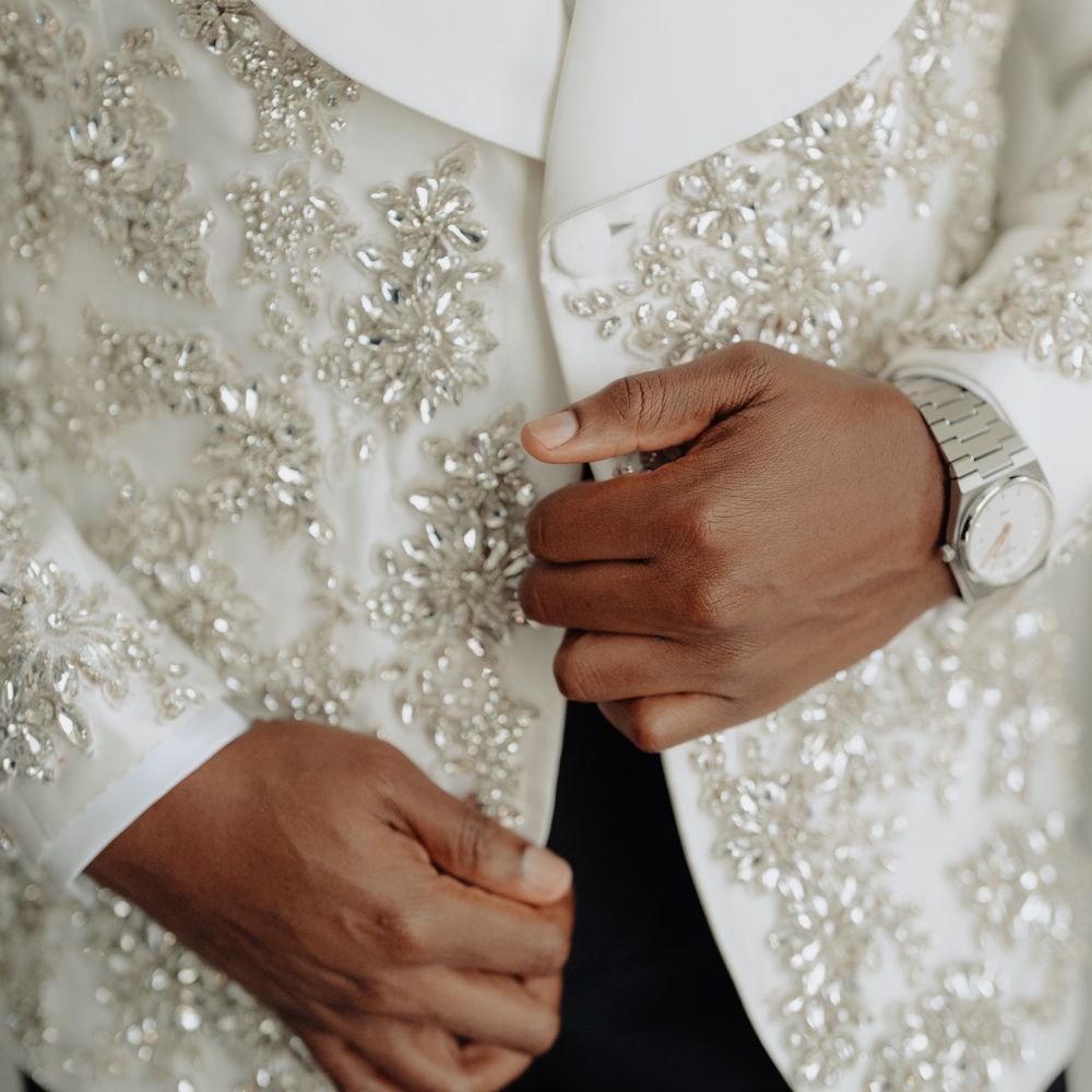 Groom in an ivory crystal-embellished tuxedo jacket adjusting his silver wristwatch