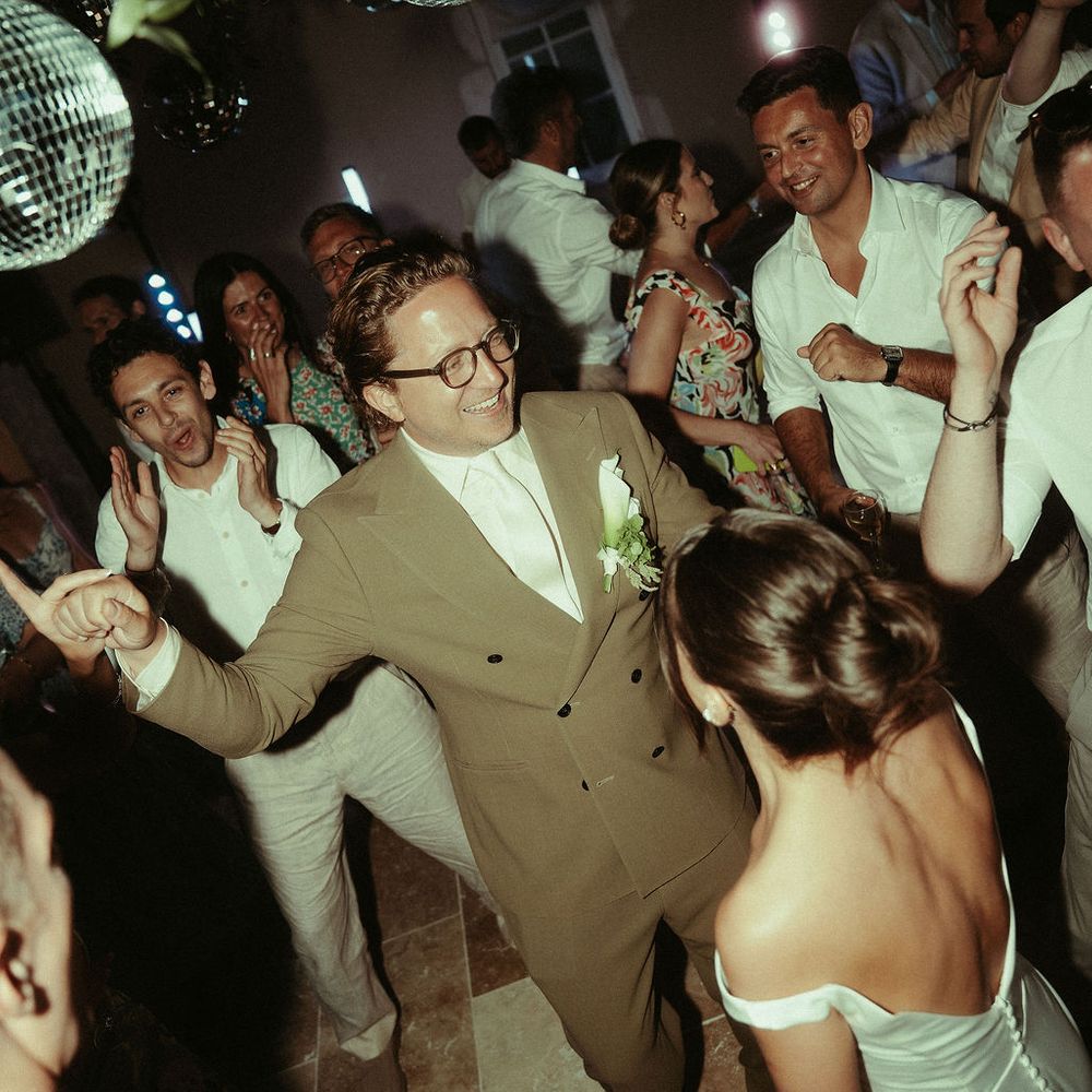 Groom in olive suit dancing with guests beneath disco balls and foliage at an evening reception