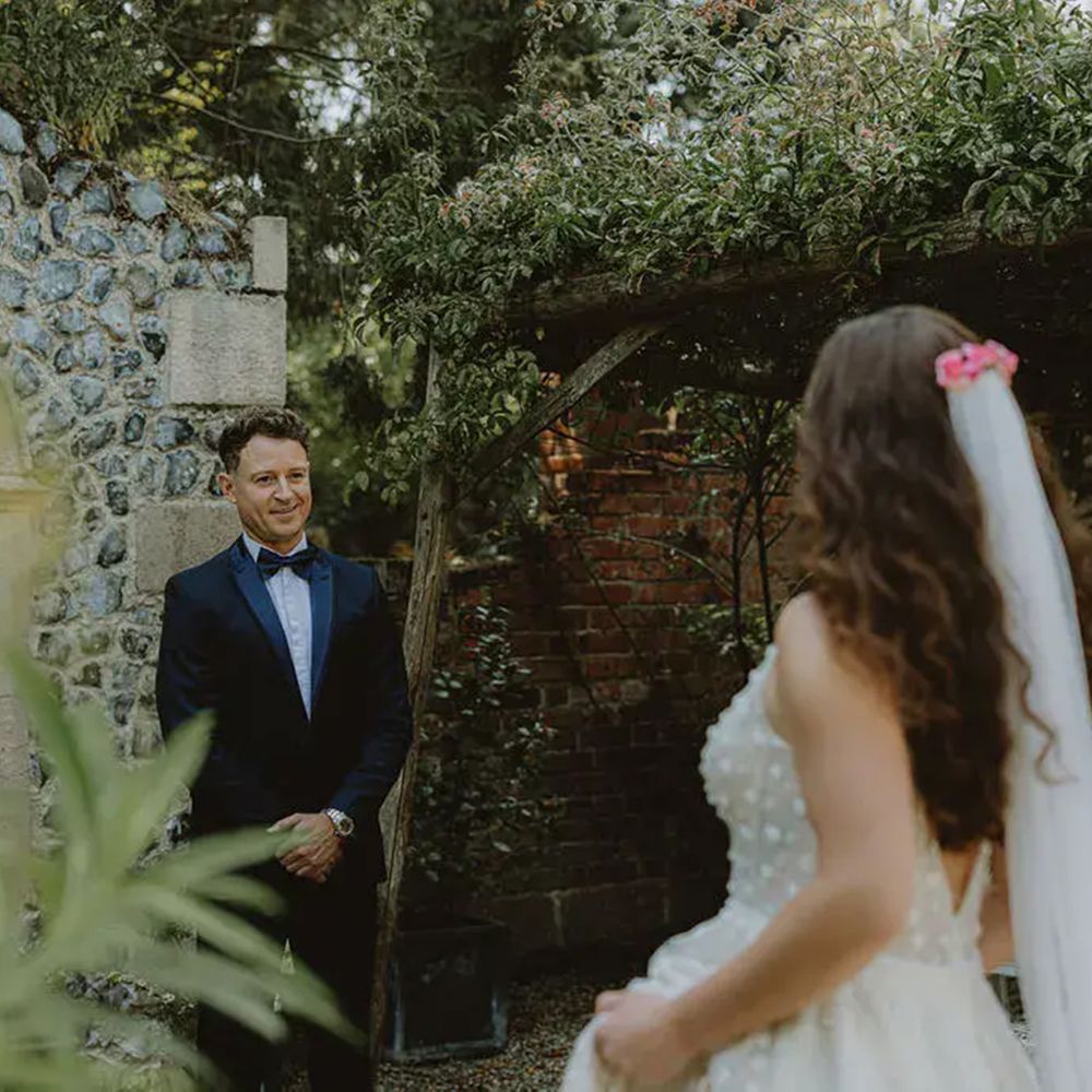 Groom in navy tuxedo smiles at approaching bride in a walled garden with flint walls