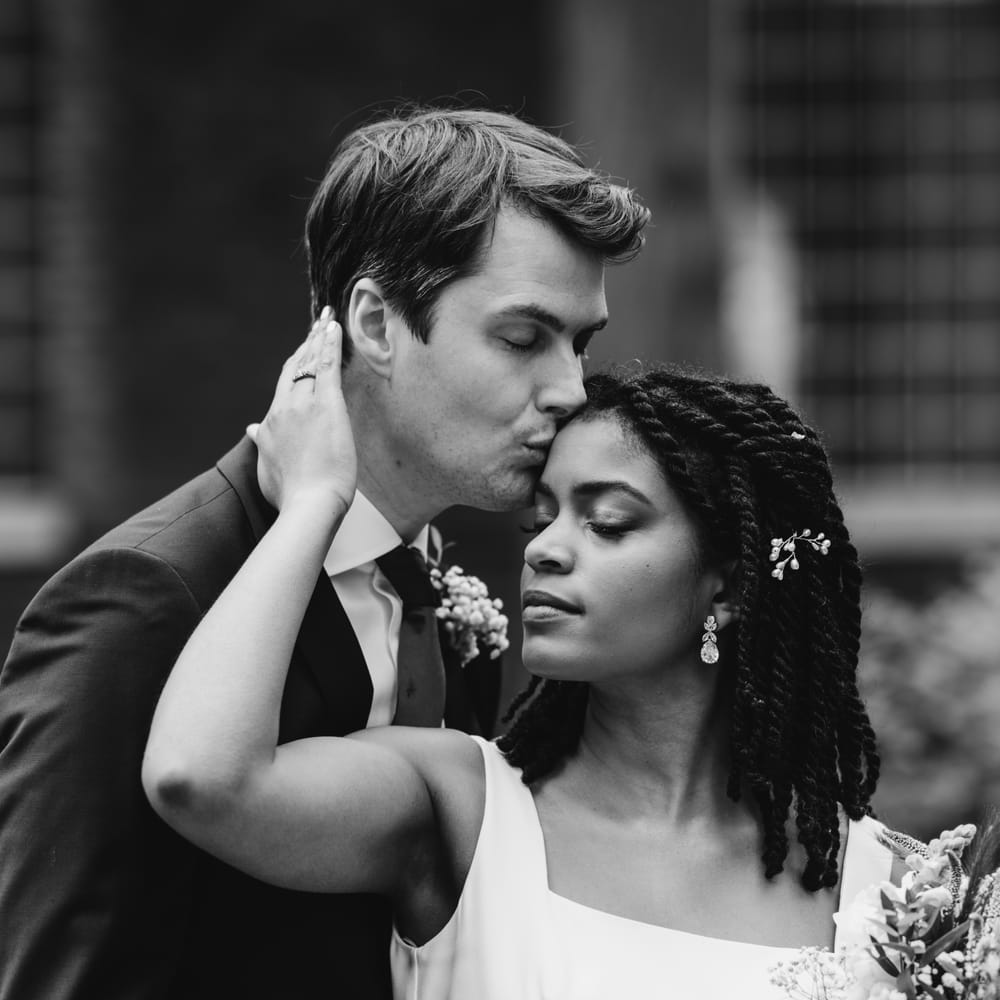 Groom kisses bride's forehead as she holds her bouquet in a tender black and white portrait