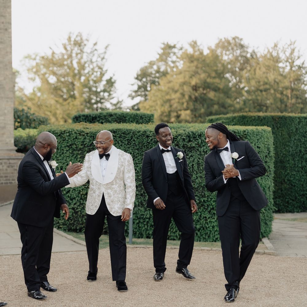 Groom in an ivory jacquard dinner jacket laughs with groomsmen in black tuxedos outside a manor