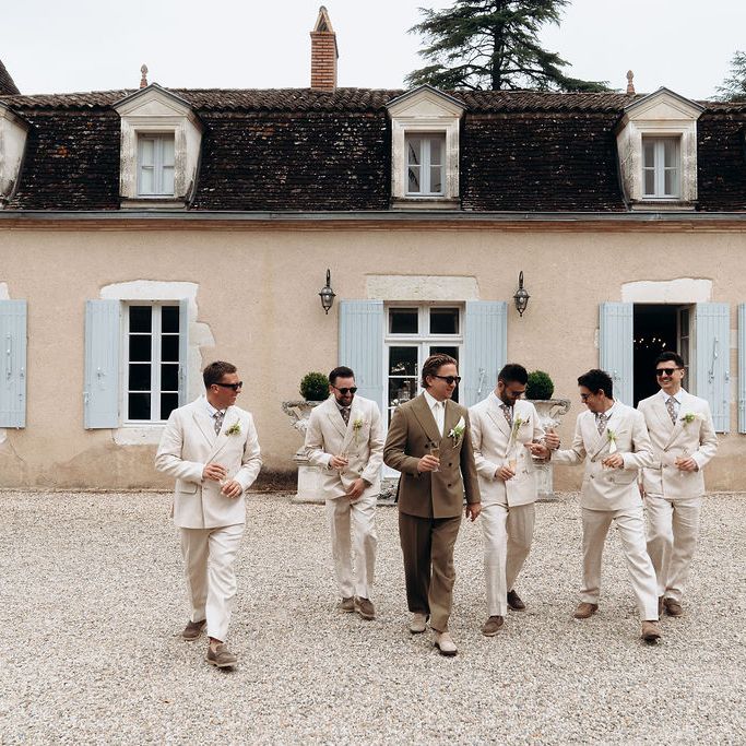 Groom in tan suit walks with groomsmen in cream suits outside a French château