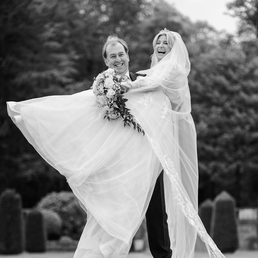 Groom lifts laughing bride whose tulle gown and veil billow out in a formal topiary garden