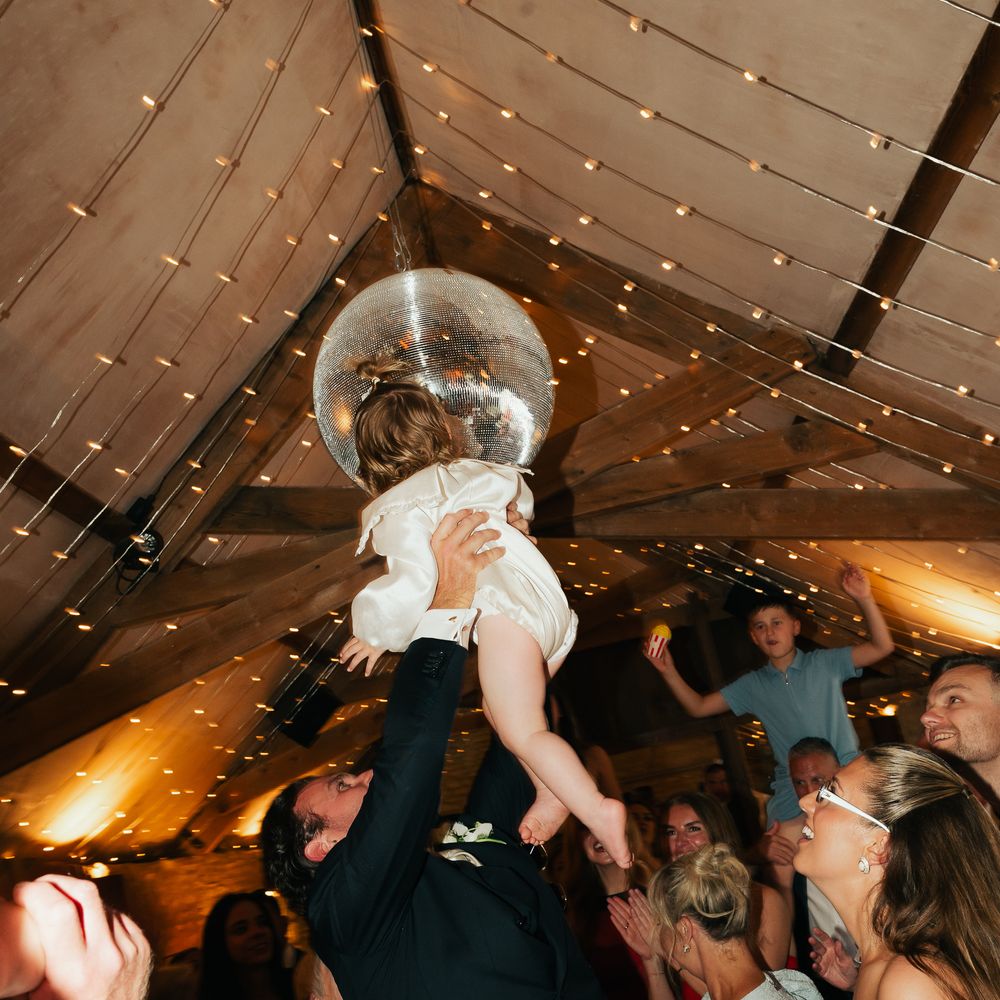 Groom lifts child towards a mirrorball beneath fairy-lit rafters as guests dance and cheer