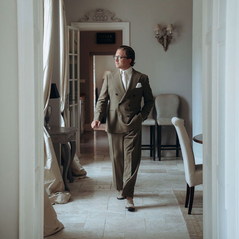 Groom in an olive double-breasted suit walking through an elegant stone-floored interior