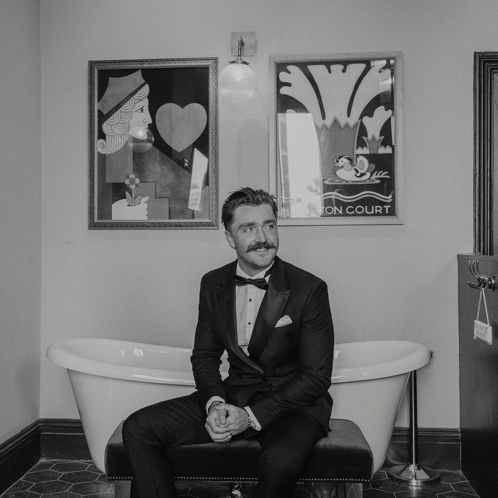 Groom in black tuxedo and patterned socks sits on a bench beside a roll-top bath