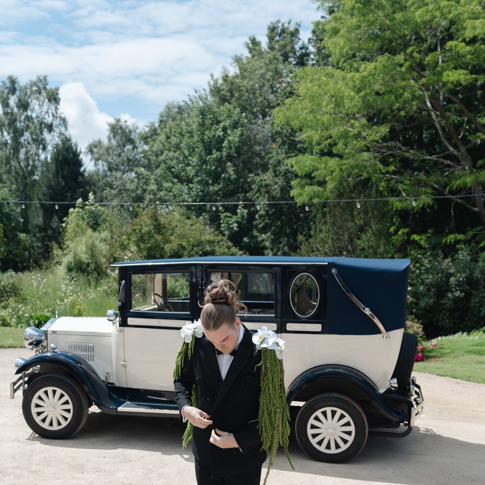 Groom in black tuxedo with cascading floral shoulder pieces beside a vintage navy and cream car