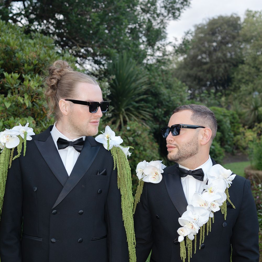 Two grooms in black tuxedos with orchid shoulder florals holding hands in a garden