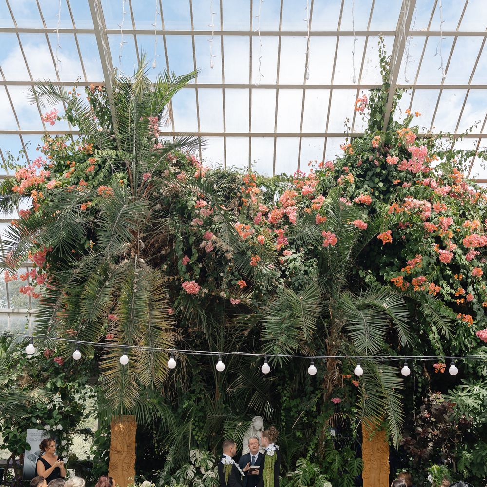 Two grooms exchange vows beneath tropical foliage and pink blooms in a glasshouse ceremony.