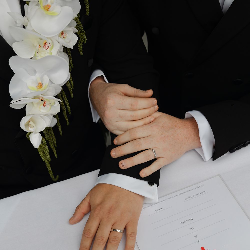 Two grooms in black tuxedos join hands showing wedding rings beside a marriage certificate.