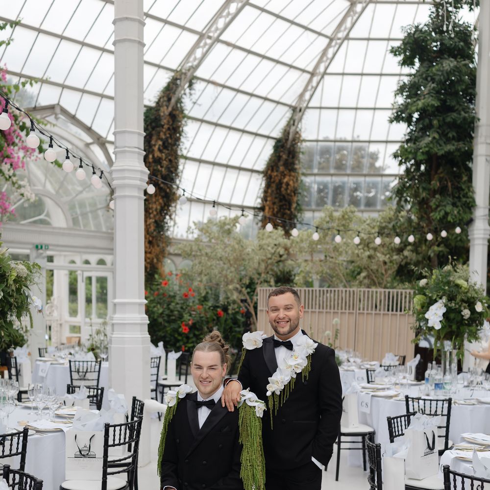 Two grooms in black tuxedos with orchid shoulder florals in a glass-domed conservatory reception