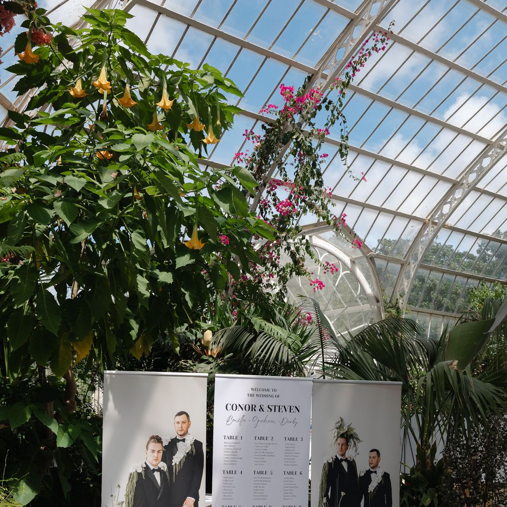 Table plan display with grooms' portraits and white florals inside a glasshouse venue.