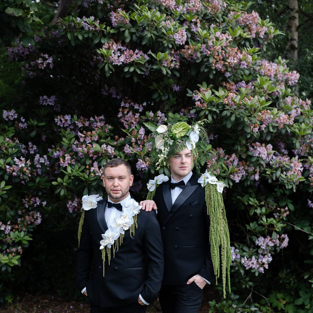 Two grooms in black tuxedos with orchid and trailing greenery details before a rhododendron bush
