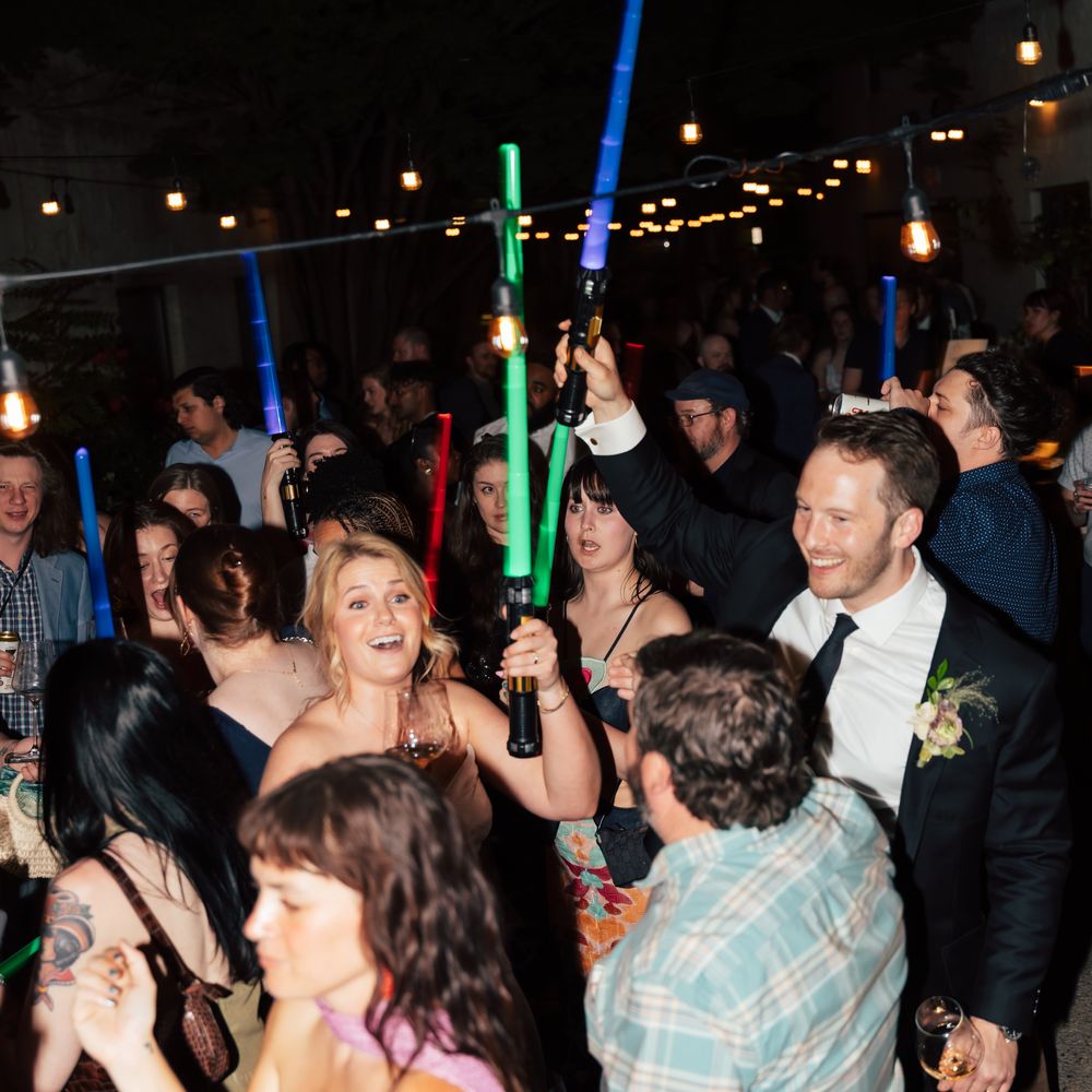 Wedding guests dancing outdoors at night holding glowing blue and green light sabers under festoon lights