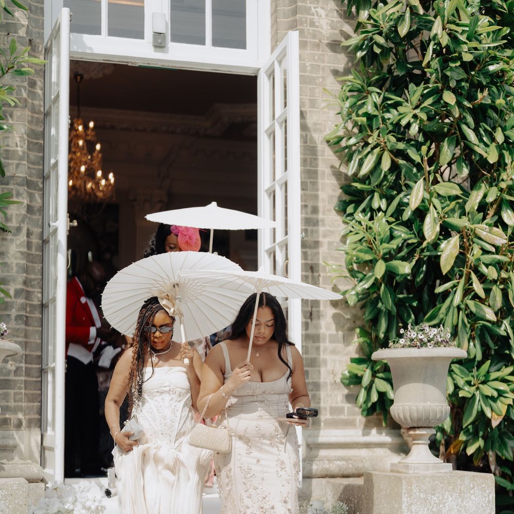 Two guests in ivory gowns carry white parasols down a floral-lined staircase from a manor house